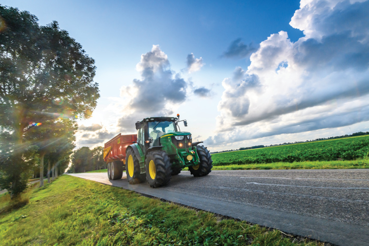 John Deere tractor hauling a loader on a country road in between agricultural fields.