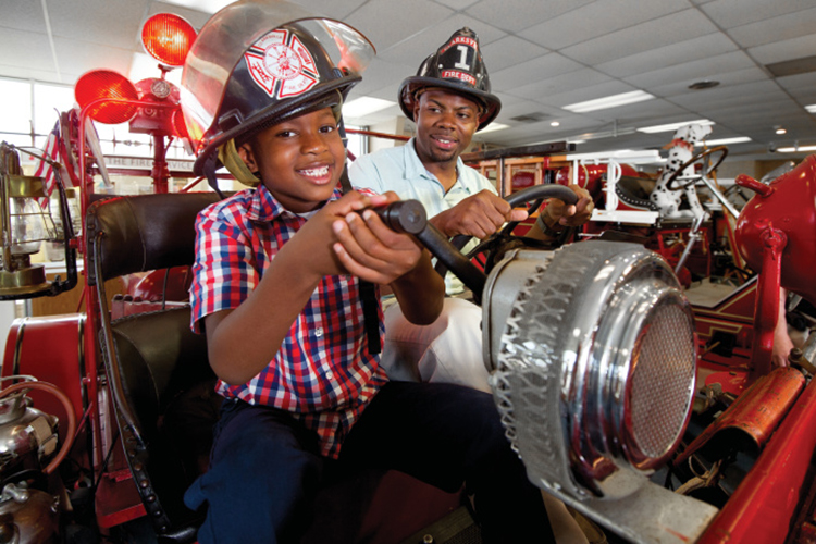 Father and son posing for a picture on a fire truck with fire hats