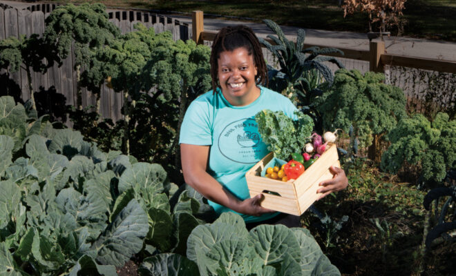 Danielle Guerin holds a box of produce standing in a field with a variety of greens growing
