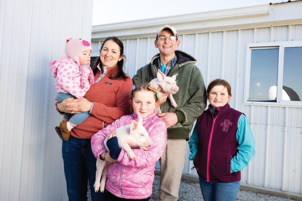 Beth holds Grace standing by Nick and Grace who are holding piglets and Hannah