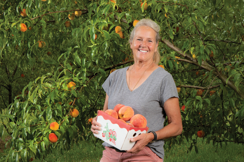 Anne Brummet holds a box of peaches with peach trees in the background at Annie's Orchard