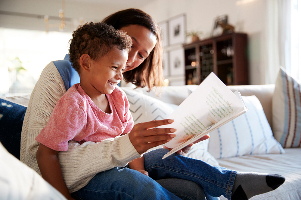 Close up of young mother sitting on a sofa in the living room reading a book with her toddler son, who is sitting on her knee, side view